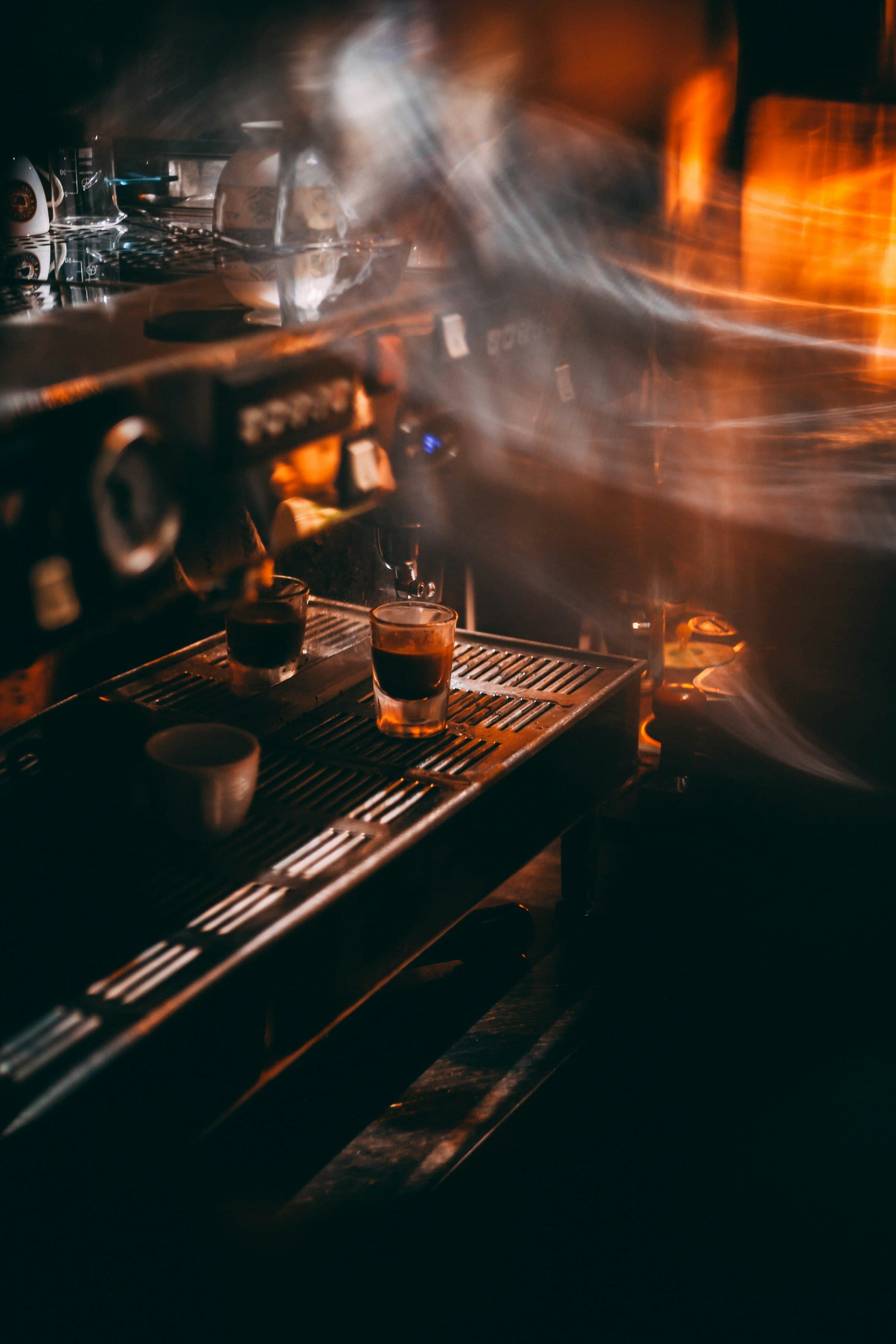 Fresh espresso in shot glasses on the lever machine's drip tray, steam drifting through amber light