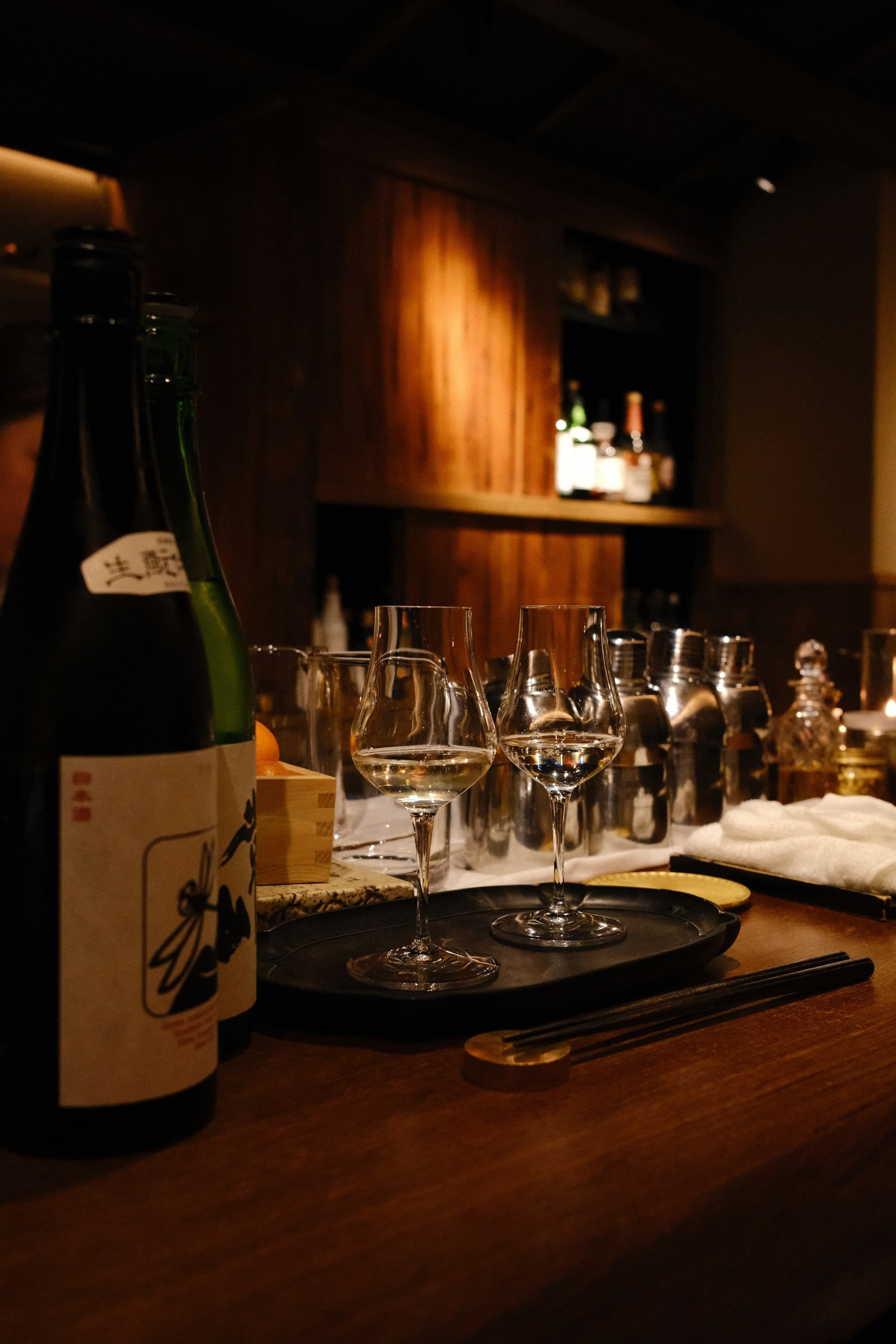 Bottles with hand-drawn labels and two tulip glasses on a black tray at the back bar